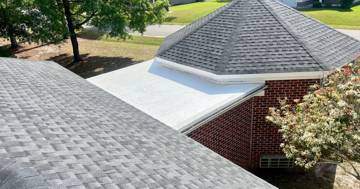 Gray architectural shingle roof meeting a white flat roof section on a brick home
